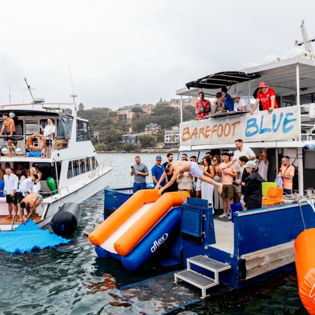 Two boats, acting as a Yacht Social Club, are docked side by side on a lake, hosting an upbeat water party. One boat named "Barefoot Blue" features two inflatable slides. Guests relish swimming, lounging, and socializing on the decks and in the water with various inflatables.