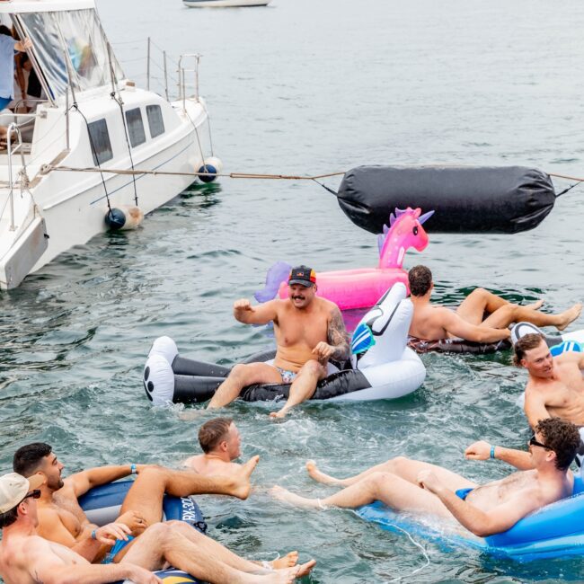 A lively social club gathers on inflatable floats, including a unicorn and a flamingo, enjoying the water near a yacht. With others in the background having fun on board, the scene is vibrant and full of joy.