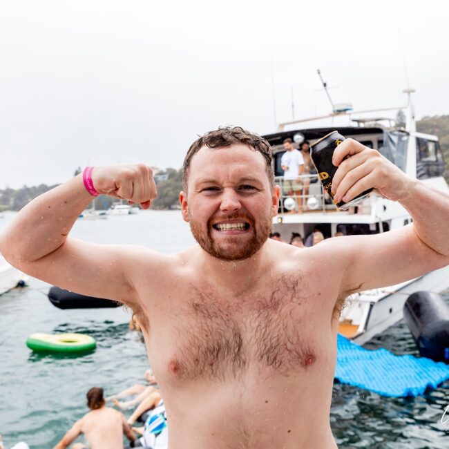 A shirtless man is flexing his muscles and holding a drink can, smiling on a yacht in a lively social club scene. He's surrounded by people relaxing on inflatables in the water, with a large boat in the background under a cloudy sky.