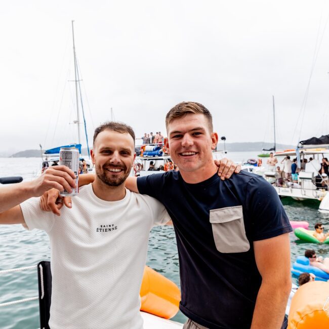 Two men smile at the camera, one holding a drink, while standing on a yacht in a marina. Several boats and people are in the background, enjoying their day at the Yacht Social Club. The weather appears overcast.