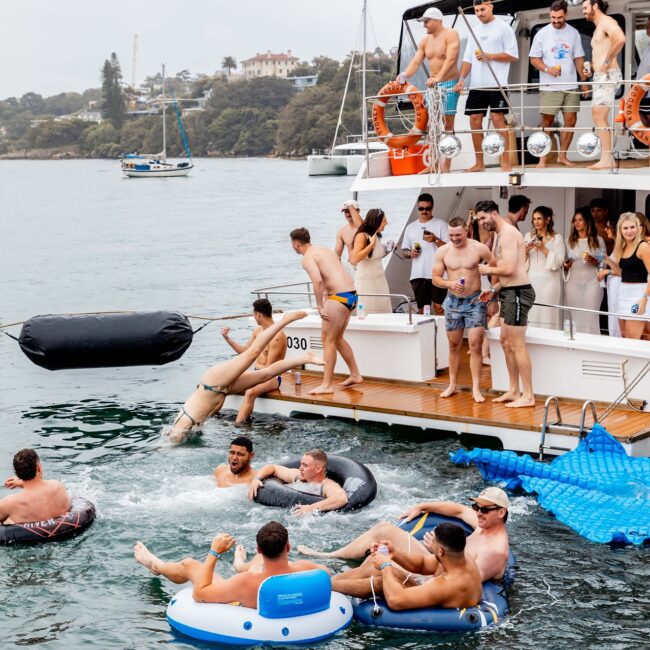 A lively social club revels on a yacht, where some members enjoy the deck's vibrant music and drinks while others float on inflatable rings. The backdrop of calm waters and distant trees adds to the festive atmosphere.