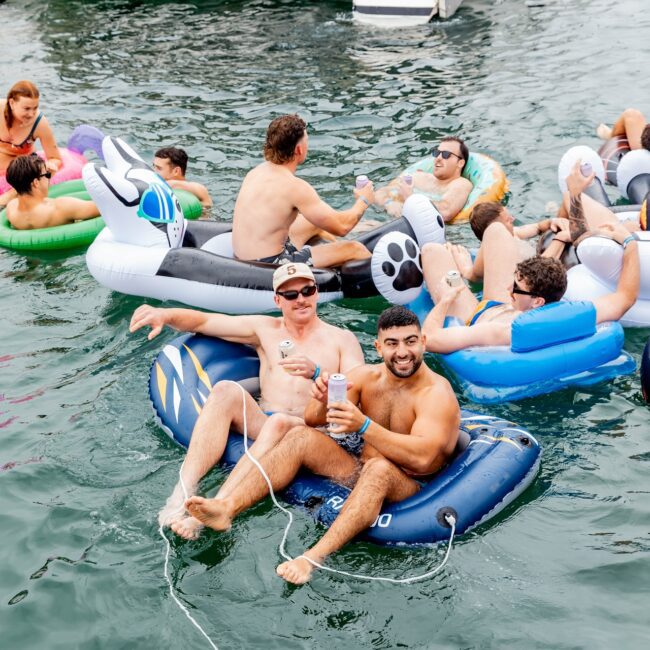 A lively social club gathers on inflatable floats near a luxurious yacht, sipping drinks and mingling under the sun. Sporting swimwear and sunglasses, they enjoy the summery vibe with boats and fellow revelers in the background.