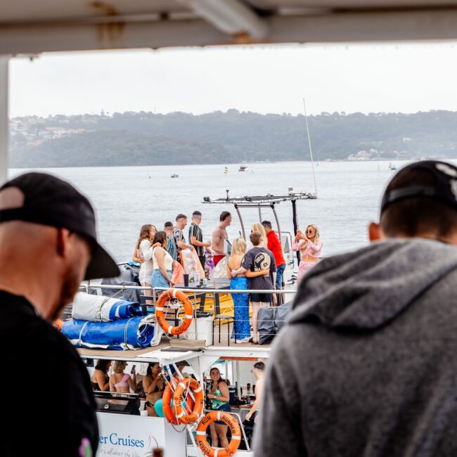 A lively group of people socializing on yachts in a scenic waterside setting. The foreground shows two club members facing away. Hills and trees line the distance under a cloudy sky, with "The Yacht Social Club" logo visible.