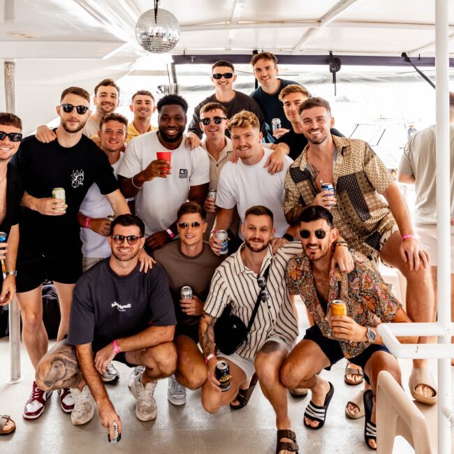 A group of sixteen men pose together on a yacht. They are smiling, dressed in casual summer attire, and holding drinks. A disco ball is visible above them. The setting appears to be bright and lively, suggesting a party hosted by their social club.