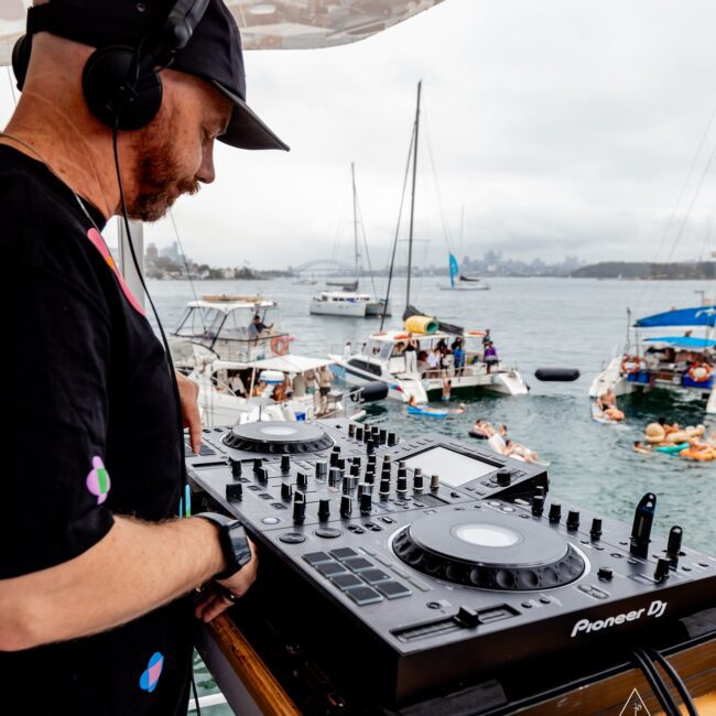 A DJ wearing a black cap and shirt is performing on a boat with a Pioneer DJ console. In the background, several people are on inflatable rafts and boats on a cloudy day. The "Yacht Social Club" logo is visible on the bottom right.