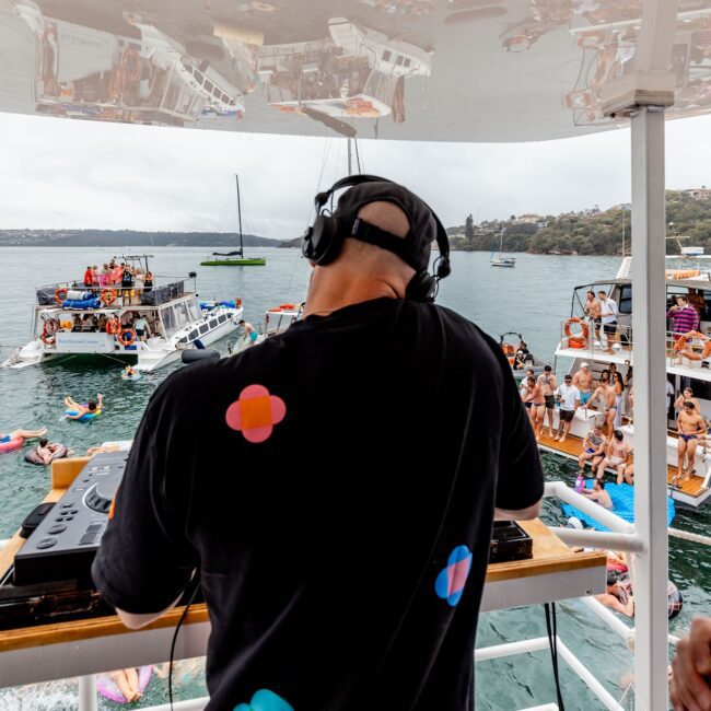 A DJ wearing headphones performs on a Yacht Social Club deck. A crowd of people on boats and inflatables enjoys the music on a sunny day by the water. The scene is lively, with reflections dancing across the ceiling above the DJ.