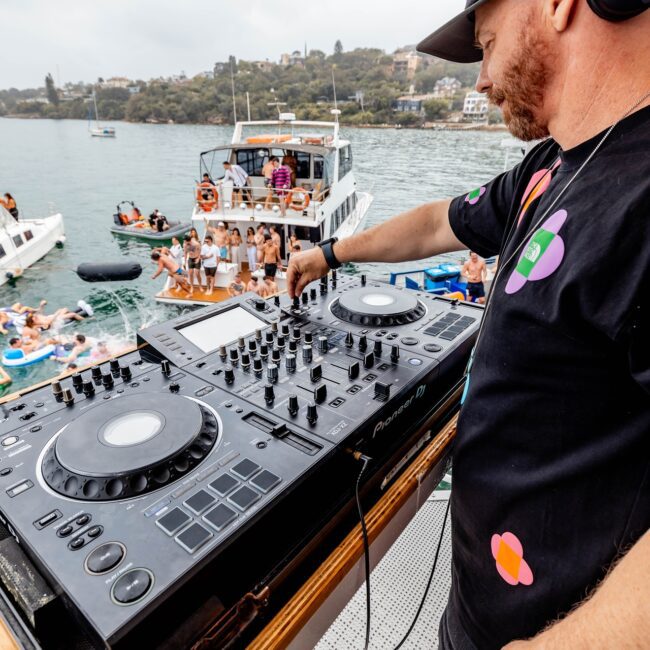 A DJ wearing headphones spins tunes on a yacht equipped with a mixer and turntables. In the background, members of a social club on a nearby boat and in the water revel in the lively atmosphere. The scene overlooks a serene body of water with distant trees and houses.