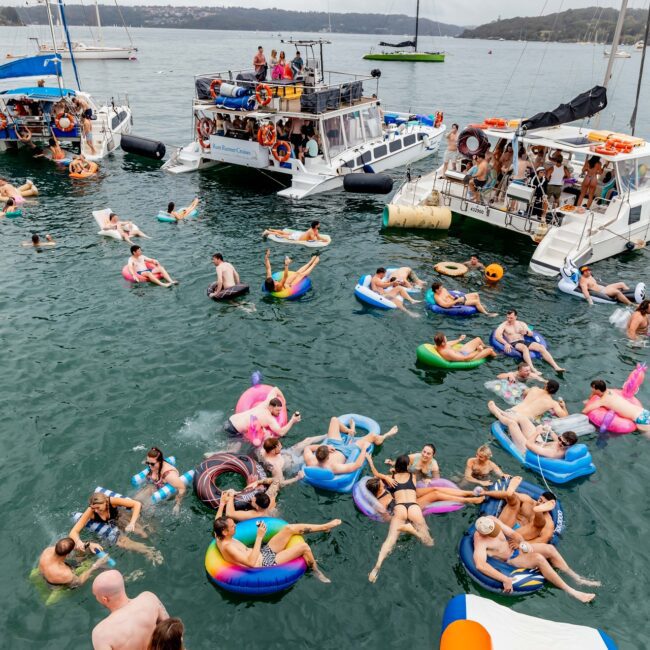 A lively outdoor party on the water with numerous people enjoying themselves on inflatable floats. Nearby yachts are anchored, exuding a social club vibe as the water is filled with people swimming and socializing under a cloudy sky.