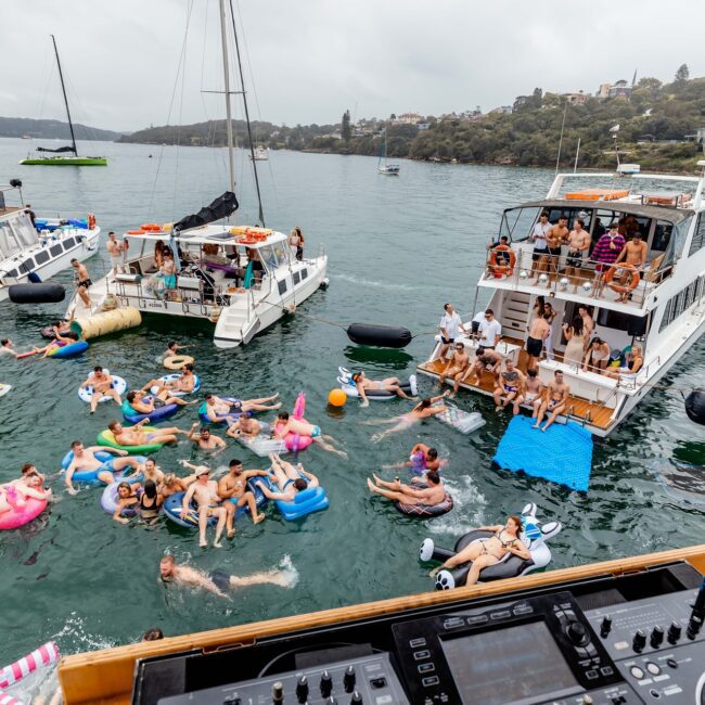 A lively yacht party hosted by the Yacht Social Club, viewed from behind a DJ booth. People are swimming and lounging on inflatables between several docked yachts. The atmosphere is festive, with a cloudy sky and a forested shoreline in the background.