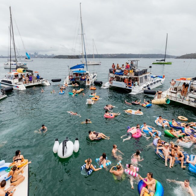 People are enjoying a social gathering on the lake, surrounded by yachts and several boats. Many are on colorful inflatable floats in the water, with more relaxing aboard. The atmosphere is lively and festive under a cloudy sky, reminiscent of an exclusive social club event.