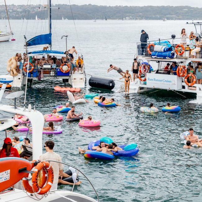 People are enjoying a lively yacht party on the water, reminiscent of a vibrant social club. Some revelers float on colorful inflatables, while others mingle on boats. The scene is festive with cloudy skies in the background.