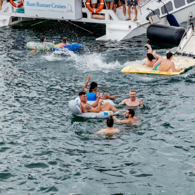 People swimming and floating on inflatables near a yacht labeled "Rum Runner Cruises" in a sparkling body of water. The boat, reminiscent of a bustling social club, has passengers on board and lifesavers attached. The scene is lively and social.