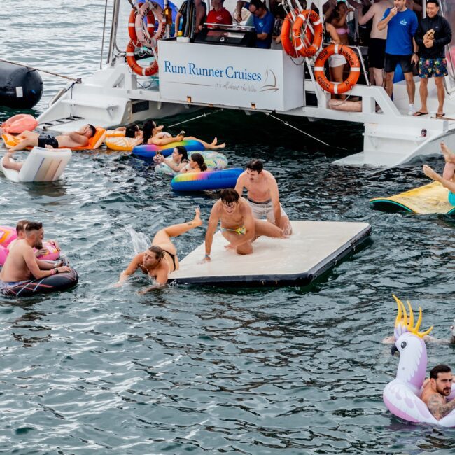 People enjoy a lively scene on the water, with some from the Social Club on a large floating platform and others on inflatables. A yacht labeled "Rum Runner Cruises" is in the background, with more people onboard. An inflatable unicorn is also visible in the water.