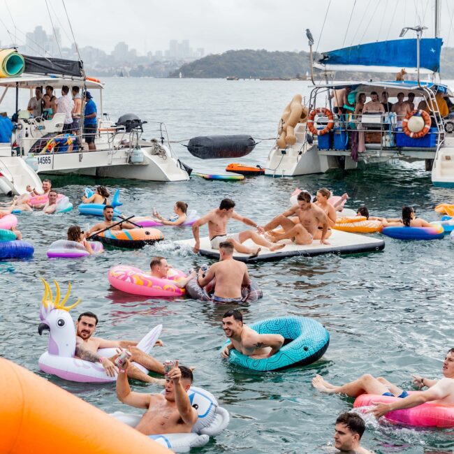 People from the social club enjoy a party on the water, floating on colorful inflatable rafts near two anchored yachts. The scene is lively with participants laughing and socializing under a cloudy sky.