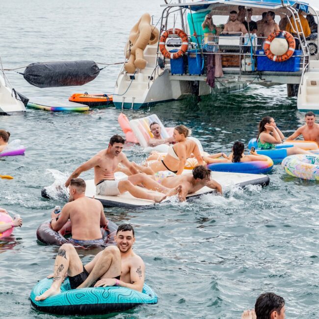 People enjoy a fun day on the water, lounging on inflatable floats near a yacht. The scene is lively, resembling a vibrant Social Club with participants splashing and socializing. The backdrop features a coastline and a touch of city skyline under a cloudy sky.