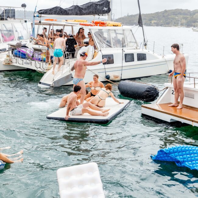 A lively scene on the water with people enjoying a yacht party unfolds like a social club gathering. Several revelers float on inflatable rafts alongside docked boats, while others mingle on the deck. Brightly-colored swimwear and a cloudy sky complete the vibrant atmosphere.