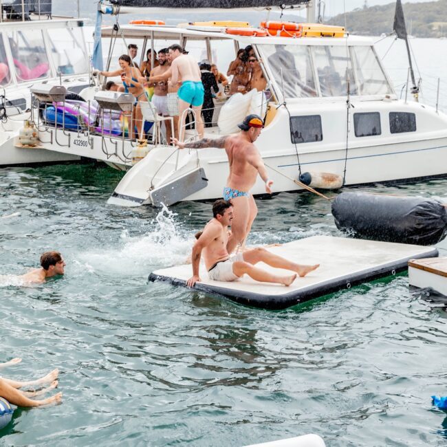 People are enjoying a lively day on the water, jumping from an inflatable mattress alongside docked yachts. Some are swimming, while others socialize aboard the vessels like a vibrant social club. The scene reflects a joyful, adventurous atmosphere.