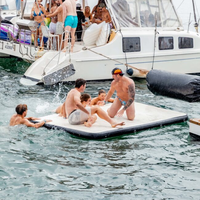 A group of people enjoying a sun-drenched day on the water, with some lounging on a floating mat and others aboard a yacht. This festive scene resembles a lively Yacht Social Club gathering, with everyone in swimwear, engaging in cheerful conversation and laughter.