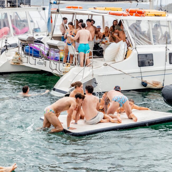 A lively and festive scene unfolds as a social club gathers in swimsuits, enjoying a party on yachts and a floating platform. Some revelers stand tall, while others splash in the water or climb onto the platform, all beneath the backdrop of cloudy skies.