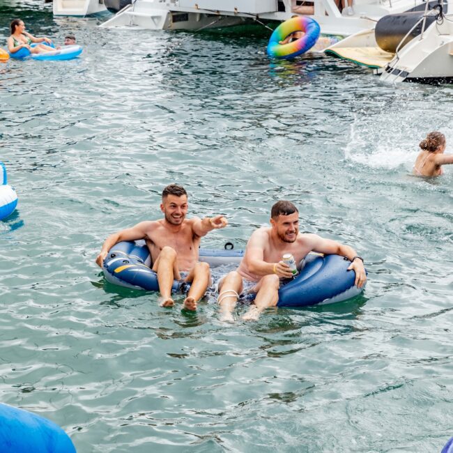 Two people happily float on inner tubes in the water, holding drinks. In the background, a yacht named "Rum Runner Cruises" is anchored with additional people onboard and in the water. The scene is lively and festive, like an exclusive social club on the waves.