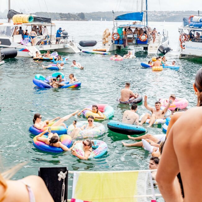 A lively party scene unfolds on the lake, with people enjoying themselves on colorful inflatable floats. Nearby, a yacht is anchored, creating a perfect backdrop as partygoers from the social club mingle and relax in the overcast waters.