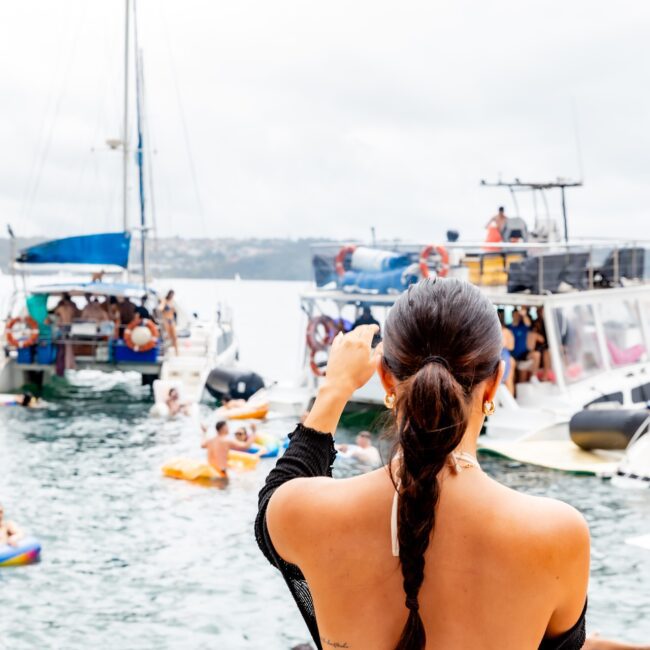 A woman with a braided ponytail gazes over the bustling waterfront, where people are swimming and relaxing on colorful floaties. Several boats from the Yacht Social Club are anchored nearby, adding to the lively scene beneath an overcast sky.