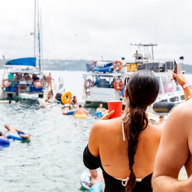 A woman in a black bikini holds a red cup and gestures towards several yachts on the water. People from the social club are relaxing on inflatable floats. A man with sunglasses and drinks is partially visible, capturing the lively essence of an outdoor yacht party.