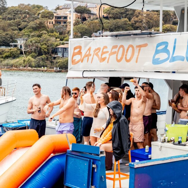 A group of people in swimwear are gathered on a yacht, enjoying a sunny day with drinks. "Barefoot Blue" is displayed on the boat's canopy. There’s a water slide in the foreground and lush greenery in the background, creating the perfect social club atmosphere.