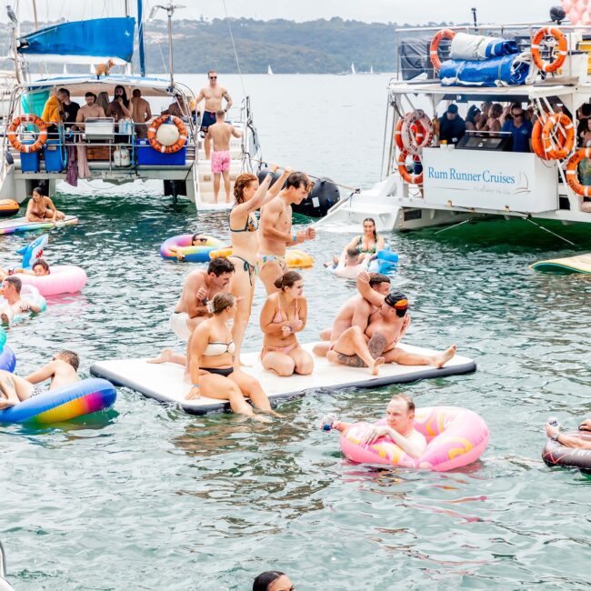 A lively scene on the lake features a party hosted by the social club. Individuals are on colorful inflatables and paddle boards, while a yacht anchors nearby. Everyone is relaxed and socializing under the cloudy sky.