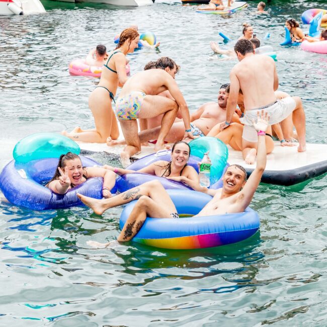 People enjoying a sunny day on the lake, floating on colorful inflatables and splashing in the water near boats. Smiling and waving, they exude relaxed cheerfulness, turning the scene into a lively Yacht Social Club celebration.