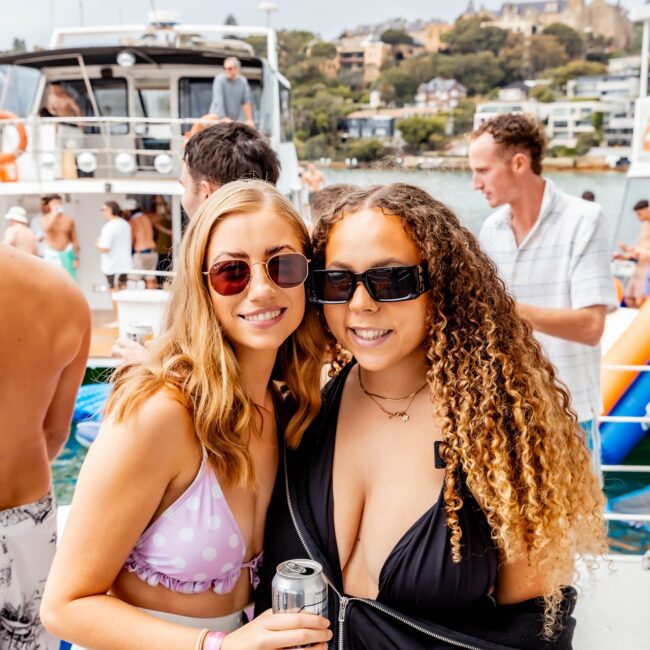 Two people smiling and holding drinks on a boat, embracing the lively atmosphere of a Yacht Social Club. Both are in sunglasses and swimwear, surrounded by others enjoying the sun. In the background, another yacht graces the scenic view of glistening water and lush trees.