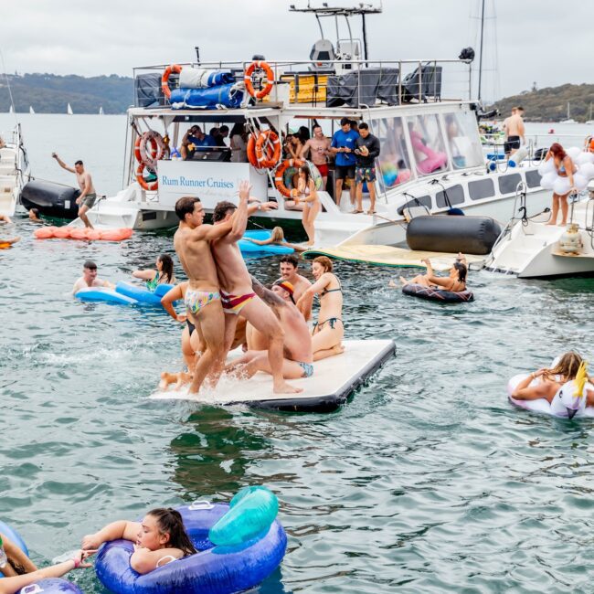 A lively scene unfolds as members of the Yacht Social Club enjoy a day on the water. Some are on inflatable tubes, while others leap from a floating platform. A yacht with life preservers and a jovial crowd on deck adds to the festive atmosphere in the background.