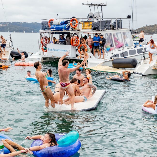 People from a social club are enjoying a lively yacht party on the water with various inflatables, including a floating mat and ring tubes. A large boat is anchored nearby, and the scene is buzzing with participants having fun under a cloudy sky.