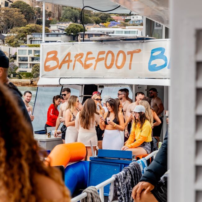 A lively group of people from a social club are enjoying a gathering on a yacht named "Barefoot Bar." They are socializing and having drinks with buildings and greenery visible in the background. Some guests are seated, while others stand around a blue counter.