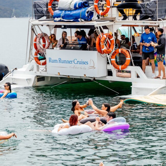 People enjoying a day on the water around a "Rum Runner Cruises" yacht. Some are on the vessel, while others float on inflatable loungers like a social club, raising drinks and mingling. The scene is lively and festive, set against a backdrop of calm waters.