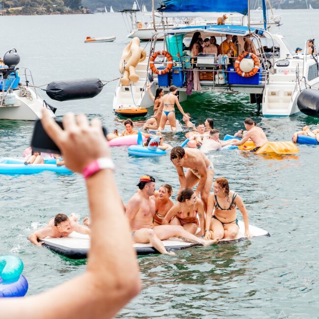 People enjoying a sunny day on the water near a yacht, with some in swimsuits on inflatable floats and others relaxing on the boat. The scene conveys a lively, social atmosphere reminiscent of a vibrant Yacht Social Club gathering in a picturesque coastal setting.