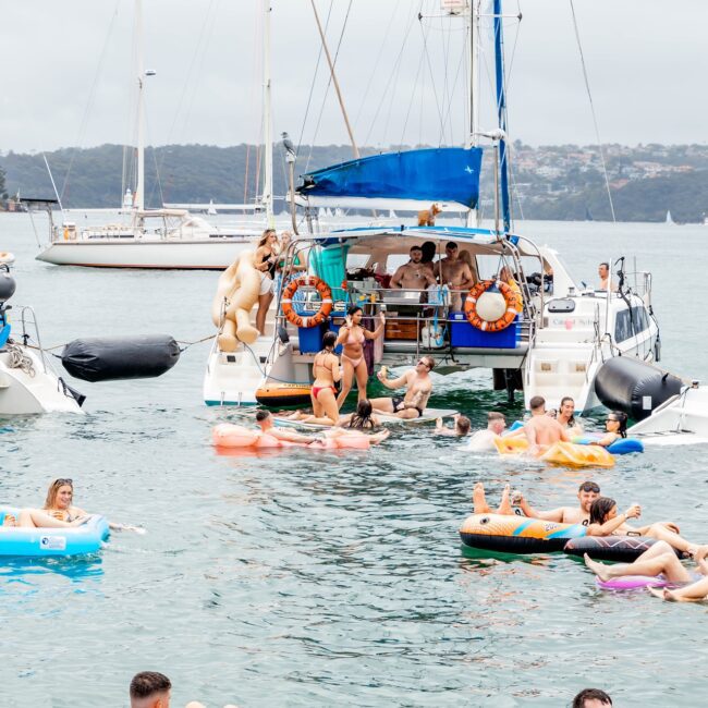 A lively social club gathers as people relax on colorful inflatable rafts in the water near a yacht. The scene is festive, with others standing or sitting on the boat. The sky is overcast, and other sailboats glide gracefully in the background.