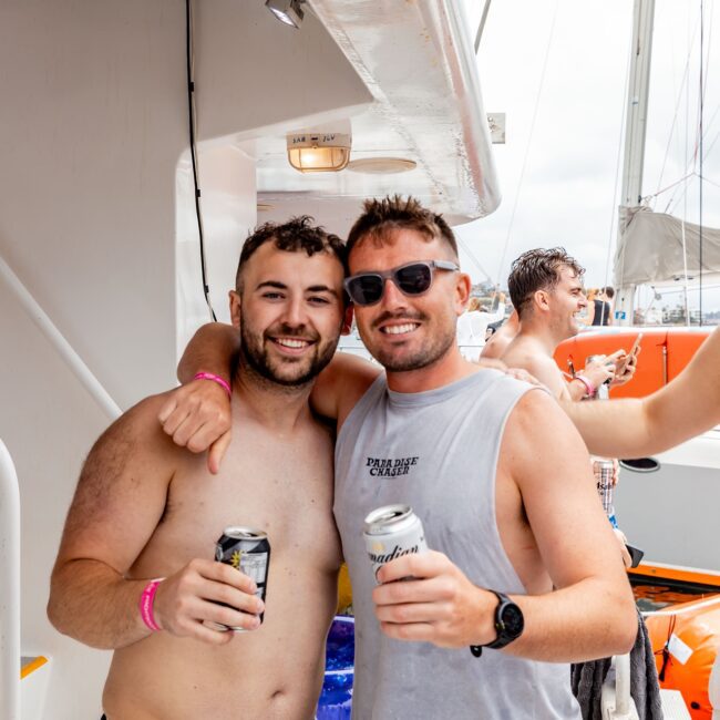 Two men with drinks pose together on a yacht, both smiling at the camera. Another shirtless man in a blue hat holds a drink in the background. The scene suggests a casual outing with the Yacht Social Club, enjoying life on the water.