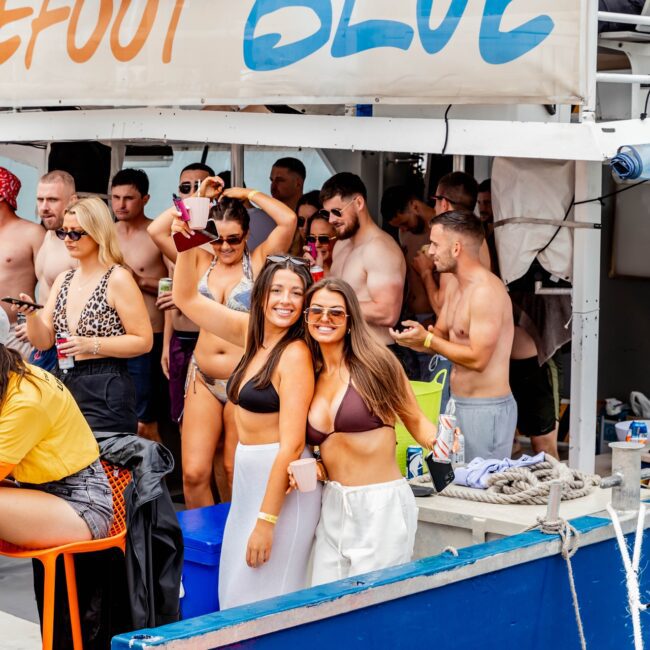 A group of people enjoying a lively yacht party. Two women in the foreground pose for a photo, smiling and wearing swimsuits. Others socialize in the background under a banner reading "Barefoot Blue," making it clear this is no ordinary gathering but an exclusive affair hosted by their social club.