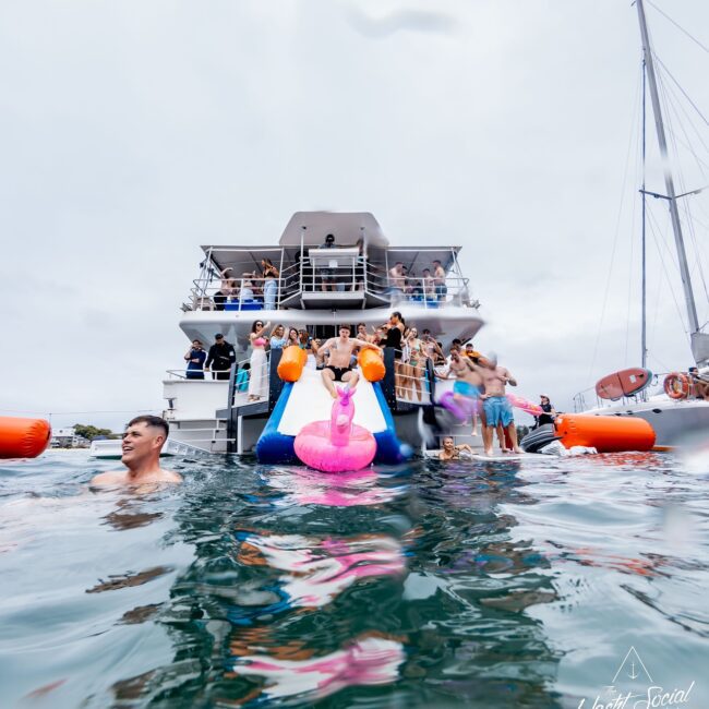 A social club revels on a yacht, with laughter echoing as members splash down the water slide. One enthusiast slides into the choppy sea on an inflatable flamingo, while a lively crowd gathers on deck, soaking in the festive atmosphere.