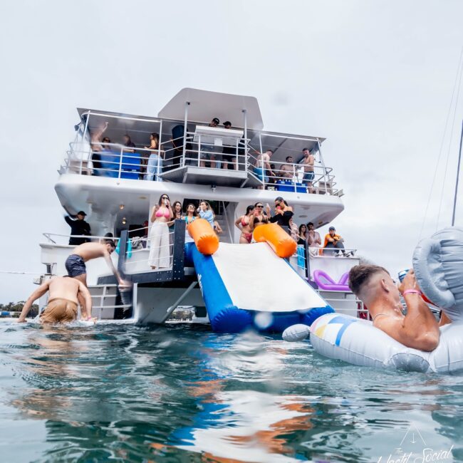 A lively party scene unfolds on a large yacht, resembling an exclusive club. Guests enjoy various activities, from gliding down an inflatable slide into the water to lounging on pool floats. With multiple decks, the yacht buzzes with social interactions and a vibrant atmosphere.