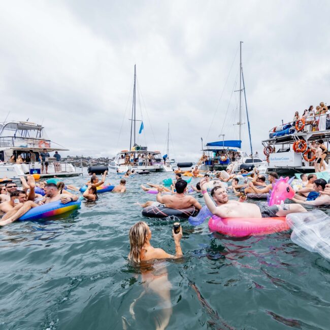 A lively Yacht Social Club party on the water features people on inflatables and yachts, enjoying drinks, music, and socializing under a cloudy sky. The scene is festive and bustling with summer vibes.