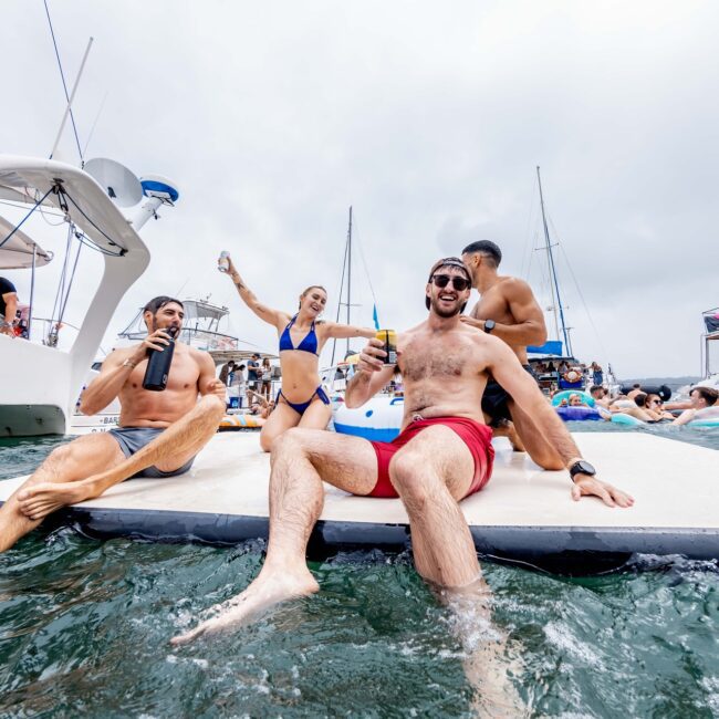 A group of friends relaxes on a floating platform in the water, with sleek yachts and fellow Social Club members in the background. They are wearing swimwear, holding drinks, and smiling at the camera, enjoying a sunny day at a lively party scene.