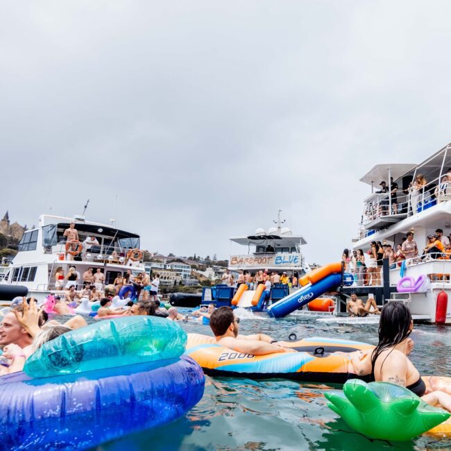 A lively scene of people enjoying a boat party on a lake, akin to a vibrant social club. Numerous individuals are on colorful inflatables in the water, surrounded by two large yachts. The overcast sky adds a casual vibe to the festive gathering.