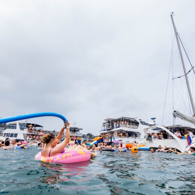 People enjoy a lively party on the water, with sailboats and yachts setting a stunning backdrop. In the foreground, a person in a pink inflatable holds a large blue noodle. This vibrant scene is reminiscent of an exclusive Yacht Social Club gathering, filled with swimming and socializing.