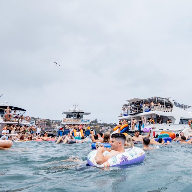 A lively scene unfolds in the bay as members of the Yacht Social Club swim and float amidst colorful floats and rings. Several boats, including elegant yachts, surround them, creating a vibrant social atmosphere even under the overcast sky. Some guests are seen mingling on the decks.