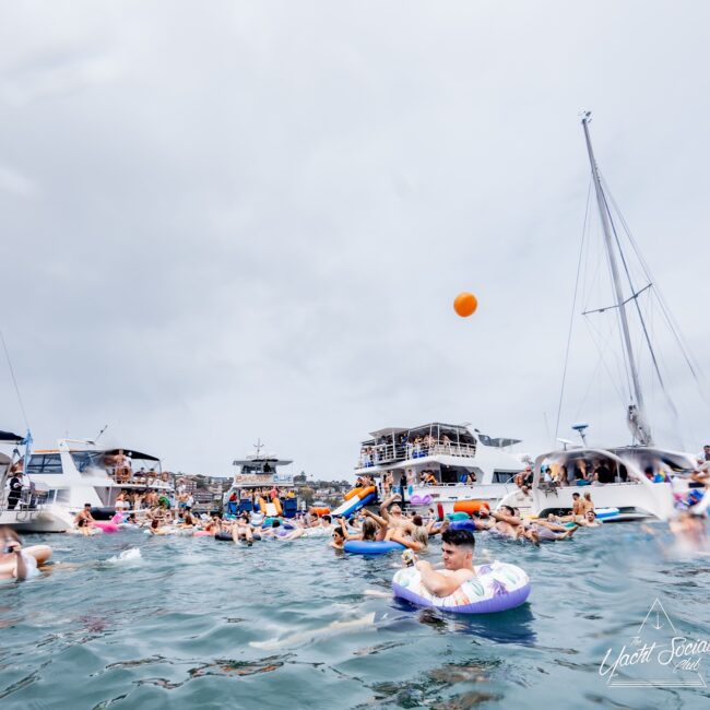 A crowded scene of people enjoying a day at sea, with numerous yachts docked closely together like a lively social club. Individuals are swimming and floating on inflatable tubes as someone tosses an orange ball amid the gathering. The sky is overcast.