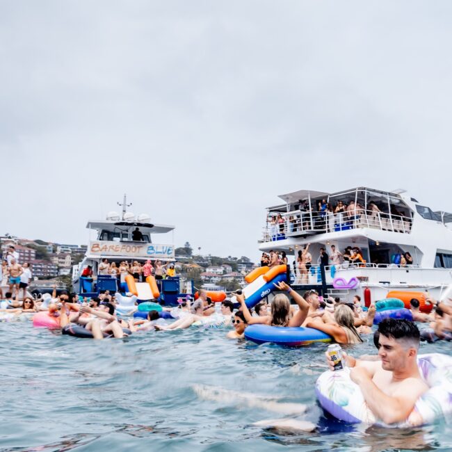 A lively scene unfolds as a Social Club revels in a day on the water. Many float on colorful inflatables, while others enjoy the luxury of a yacht. In the background, more club members are socializing on another boat, creating an atmosphere that's as vibrant as it is fun.