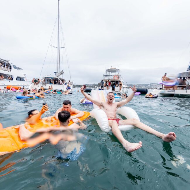 People relaxing on inflatable floats in a body of water, surrounded by anchored yachts. A man in front appears cheerful on a white float. Others are on an orange float nearby. Overcast sky and party atmosphere with music and socializing like a lively Social Club. Logo at the bottom right.