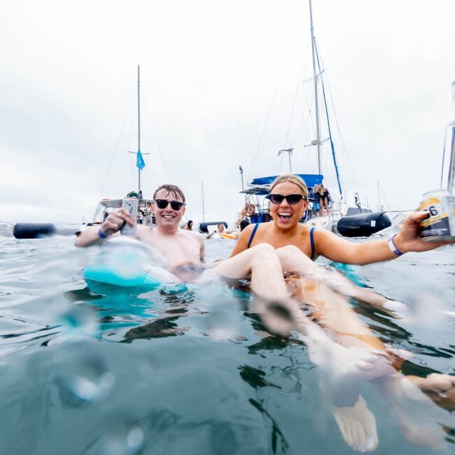 A group of friends from the social club are enjoying a day on the water, sitting on floaties and holding canned drinks. Surrounded by yachts, they seem to be having a relaxing time under a cloudy sky.